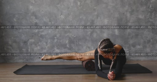 A middle-aged athletic woman performs myofascial massage of the leg muscles, rolls the muscles of the inner thigh with a special massage roller, trains in a room near the wall. Стоковое видео, видеограф Сергей Чайко / Фотобанк Лори