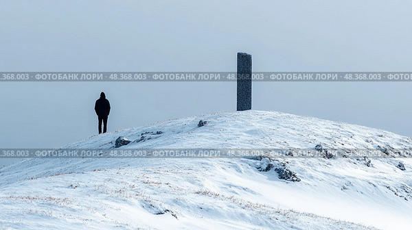 A person dressed in dark clothing stands alone on a snowy hill beside a tall monolith, surrounded by a bleak, gray sky. Стоковое AI-изображение, AI-художник Данила Большаков / Фотобанк Лори