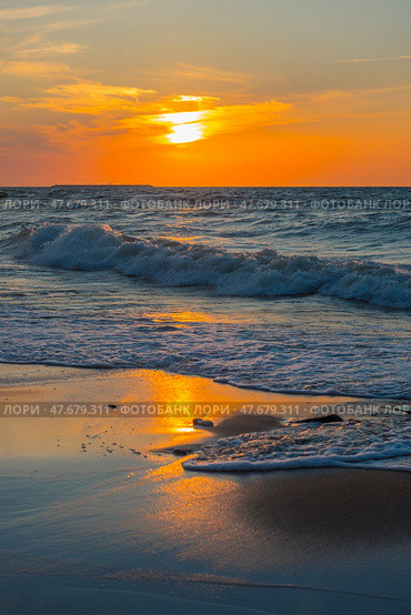 A picturesque sunset over the Baltic Sea. The sun slowly sinks below the horizon, painting the sky and water in rich shades of orange, yellow and blue. The waves gently roll onto the sandy shore, reflecting the golden light. In the foreground are wet sand covered with surf foam and wooden riots extending into the water. The atmosphere of calm, solitude and natural harmony makes the scene ideal for illustrating themes: relaxation, nature, the Baltic, meditation, travel. Стоковое фото, фотограф Анна Кривицкая / Фотобанк Лори