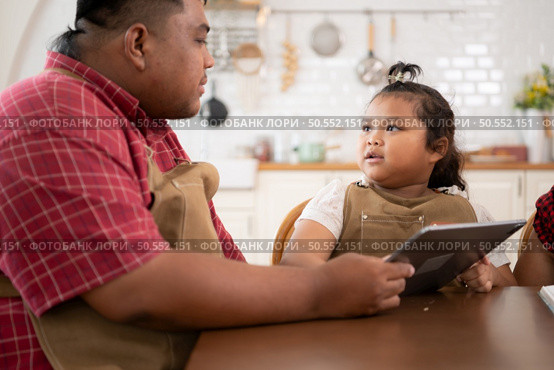 A plus-size family with a father wearing a prosthetic leg, is happily assisting a child with her homework and having fun together in the dining room of the house before cooking together. Стоковое фото, агентство Ingram Publishing / Фотобанк Лори