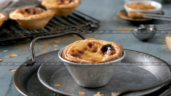 A Portuguese custard tart in a metal tin sits on a round tray with others on a rack cinnamon and measuring spoons in the background. Стоковое фото, агентство Ingram Publishing / Фотобанк Лори