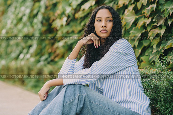 A reflective young woman in a striped shirt and jeans sits peacefully outdoors, framed by vibrant greenery, embodying tranquility and deep introspection that resonates with her surroundings. A young woman poses gracefully outdoors, surrounded by lush foliage, enjoying the serene moment. Стоковое фото, агентство Ingram Publishing / Фотобанк Лори