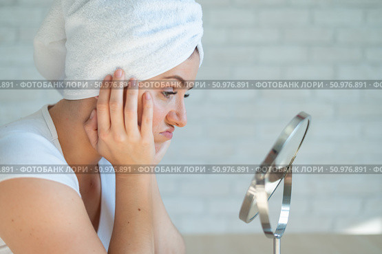 A sad woman sits at a table and looks in the mirror. A miscarriage of a girl with hormonal problems induces beauty after a shower. Teen acne. Himself a dermatologist. Problem skin. Стоковое фото, фотограф Михаил Решетников / Фотобанк Лори