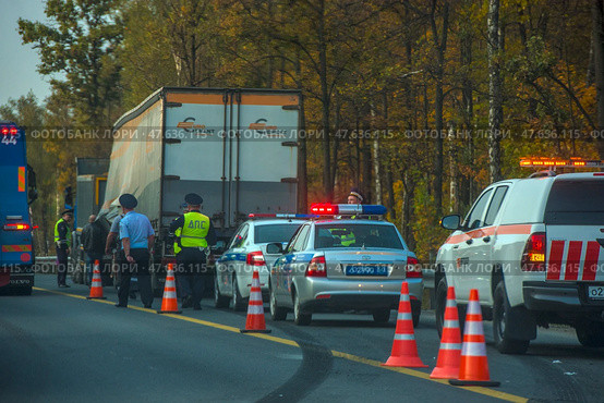 A section of road where police and road service officers are conducting an inspection or processing an incident. Several cars with flashing lights, including traffic police patrol cars and a road service vehicle, are parked on the side of the road. The road is partially blocked by orange cones. A truck with the Eurocement logo is visible in the background. In the foreground are police officers in uniform and reflective vests. The action takes place against the backdrop of an autumn forest. (2020 год). Редакционное фото, фотограф Анна Кривицкая / Фотобанк Лори