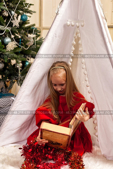 A six-year-old girl model is delightedly discovering the world within a storybook. She sits inside a charming tent surrounded by festive decorations, immersed in her imaginative adventure. Стоковое фото, фотограф Aleksandr Golubev / Фотобанк Лори