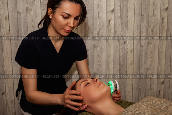 A skilled therapist performs a rejuvenating facial treatment on a client in a tranquil indoor spa. Soft light filters through the wood-paneled walls, enhancing the calming atmosphere. Стоковое фото, фотограф Aleksandr Golubev / Фотобанк Лори