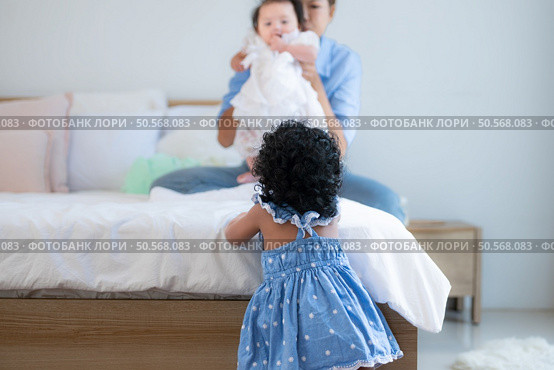 A small girl stood at the foot of the bed playing with the younger sibling carried by his mother and smiling with affection, In a bedroom surrounded by warmth and family. Стоковое фото, агентство Ingram Publishing / Фотобанк Лори