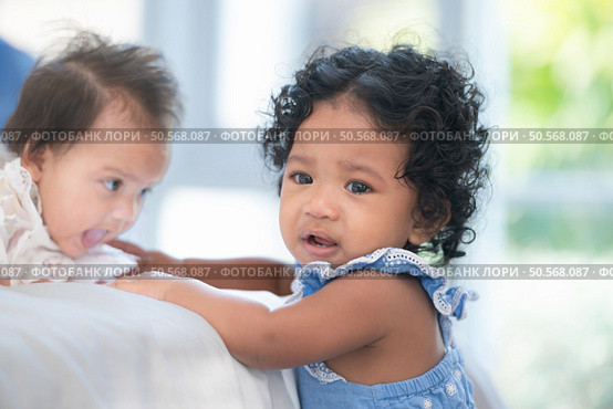 A small girl stood at the foot of the bed playing with the younger sibling carried by his mother and smiling with affection, In a bedroom surrounded by warmth and family. Стоковое фото, агентство Ingram Publishing / Фотобанк Лори