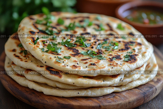 A stack of flatbreads cooked on a griddle or grill. They are garnished with fresh herbs, perhaps cilantro or parsley, adding color to the dish. The flatbreads have a golden, lightly toasted crust with characteristic spots indicating even cooking. The background is complemented by the blurry details of the greenery and perhaps sauces or dips in ceramic bowls, creating a homey or ethnic atmosphere. Стоковое AI-изображение, AI-художник Анна Кривицкая / Фотобанк Лори