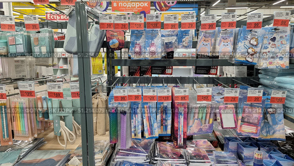 A store rack with a variety of office supplies. Sets of pens, pencils, markers, rulers, sharpeners, scissors, glue, corrector, notepads, stickers and decorative stickers with images of cute animals and cartoon characters are neatly hung on the shelves and hooks. The products are bright, colorful, and come in attractive packaging. Each product has price tags with promotional offers. In the center of the composition, colorful sets of stationery in pink, purple and blue tones stand out. The atmosphere of the photo conveys the setting of a supermarket or hypermarket with a wide selection of school and office supplies. Редакционное фото, фотограф Анна Кривицкая / Фотобанк Лори