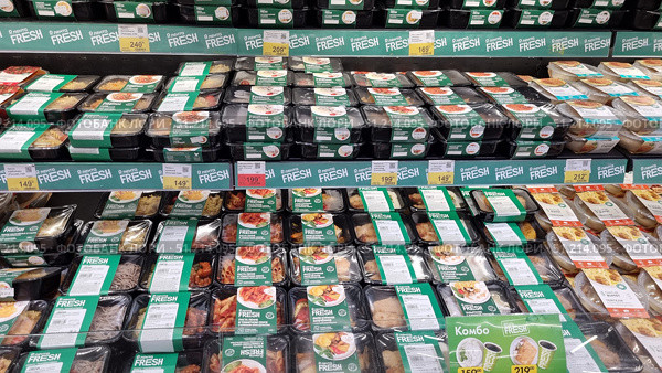 A store shelf with a large assortment of ready-made meals in packages under the Lenta Fresh brand. The products are presented in black plastic containers with a transparent lid and green labels. Various dishes are visible: pasta, meat cutlets, chicken with a side dish, noodles, vegetable stews, casseroles and other options. The packages are neatly laid out in rows on several shelves. The shelves also have price tags with promotional offers and discounts. The atmosphere of the photo emphasizes the convenience and variety of ready-made food intended for fast food at home or at work. Редакционное фото, фотограф Анна Кривицкая / Фотобанк Лори