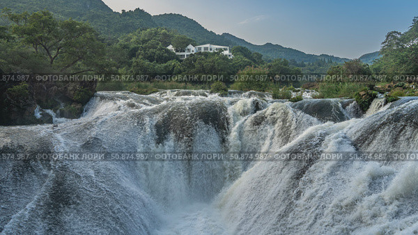 A stormy mountain river. Streams of white water are bubbling and foaming on boulders in the riverbed. Green vegetation on the shores. A building on a hill. China. Silver chain falls. Guizhou. Стоковое фото, фотограф Вера Ивановна Кубарева / Фотобанк Лори