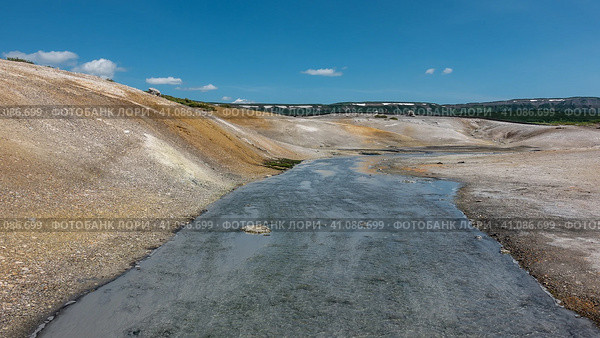 A stream from hot springs flows along a rocky bed. Yellow sulphurous deposits are visible on the soil. A mountain range against a blue sky. Kamchatka. The caldera of the extinct volcano Uzon. Стоковое фото, фотограф Вера Ивановна Кубарева / Фотобанк Лори