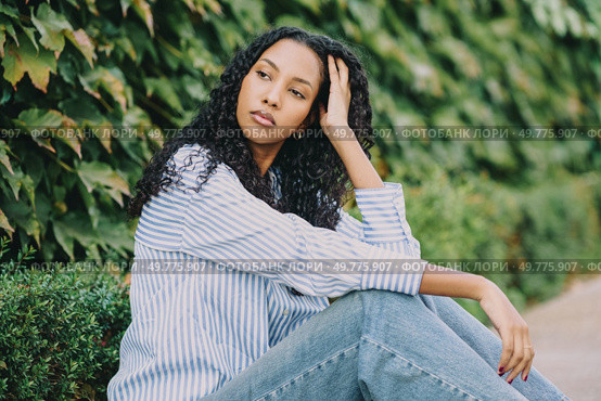 A stylish young woman wearing a chic striped shirt poses thoughtfully in an outdoor setting that is beautifully surrounded by lush green foliage, radiating both elegance and tranquility. A contemplative young woman stands against a lush green ivy wall, surrounded by nature. Стоковое фото, агентство Ingram Publishing / Фотобанк Лори