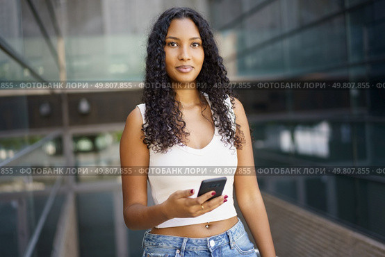 A trendy and stylish young woman dressed in a casual outfit stands confidently in an urban environment. She is holding her smartphone and directing her gaze towards the camera, exuding selfassurance. A Confident Young Woman Holding a Smartphone in a Vibrant Urban Environment, Radiating Energy. Стоковое фото, агентство Ingram Publishing / Фотобанк Лори