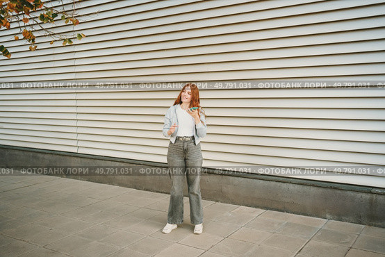 A trendy, fashionable young woman with vibrant red hair stands confidently against a contemporary urban backdrop, showcasing her distinctive and unique fashion sense with great style and flair. A stylish young woman confidently poses against a backdrop of modern architecture. Стоковое фото, агентство Ingram Publishing / Фотобанк Лори