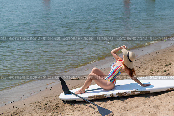 A woman in a swimsuit and a hat is posing on a sup board on the beach. Стоковое фото, фотограф Михаил Решетников / Фотобанк Лори