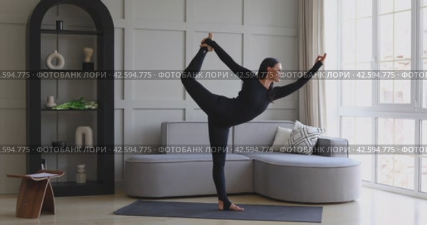 A woman leading a healthy lifestyle performs the Natarajasana exercise at home, king of the dance pose, training in a black sports overalls standing on the mat, full-length view. Стоковое видео, видеограф Сергей Чайко / Фотобанк Лори