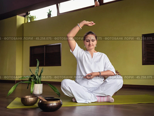 A woman practices meditation in Kundalini yoga, sits in a comfortable lotus position on a mat in a room, in white clothes, with her hands showing the Chin Mudra gesture, full-length view. Стоковое фото, фотограф Сергей Чайко / Фотобанк Лори