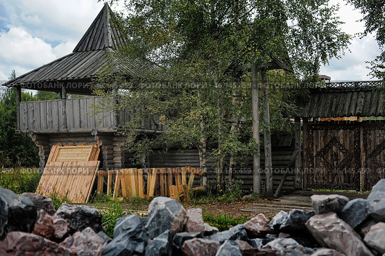 A wooden fortress or fortification built in a traditional style from logs. In the center of the frame, a massive tower with a covered terrace is visible, which can be used as a watchtower or a defensive tower. Along the perimeter, there is a wooden fence with carved gates decorated with complex geometric patterns.In the foreground, building materials are stacked - boards, beams, which may indicate the process of repair or expansion of the building. In the lower part of the frame, piles of stones are visible, possibly used for construction or protection of the territory.The surroundings consist of greenery - trees, bushes and grass, which gives the picture a natural and harmonious atmosphere. Стоковое фото, фотограф Анна Кривицкая / Фотобанк Лори