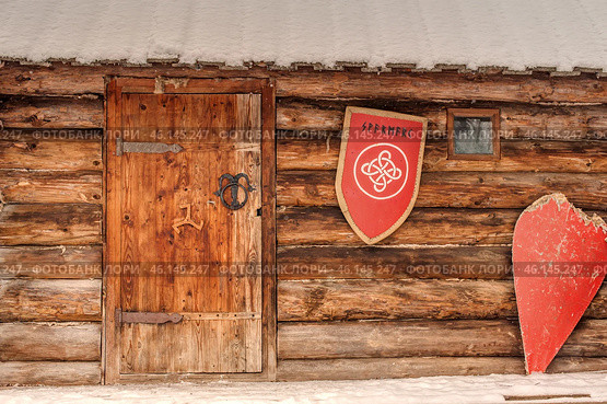 A wooden structure, possibly part of a medieval fortification or military camp. The central element is a massive wooden door with metal hinges and a forged handle decorated with symbols. A snowy roof hangs over the door. On the right side of the wall hang two red shields - one round with a coat of arms and an inscription, the other pointed, clearly battered in battle. A small square window further emphasizes the simplicity and severity of the structure. The overall atmosphere creates the impression of a reconstruction of an ancient Russian or Scandinavian settlement. Стоковое фото, фотограф Анна Кривицкая / Фотобанк Лори