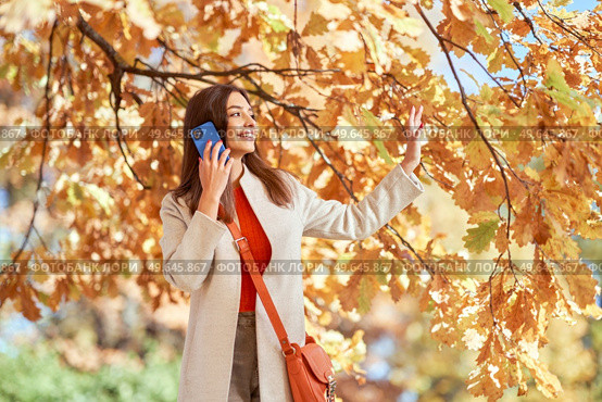 A young beautiful woman walks in the autumn park and talks on the phone. A girl with a smartphone takes photos in an autumn forest. Стоковое фото, агентство Ingram Publishing / Фотобанк Лори