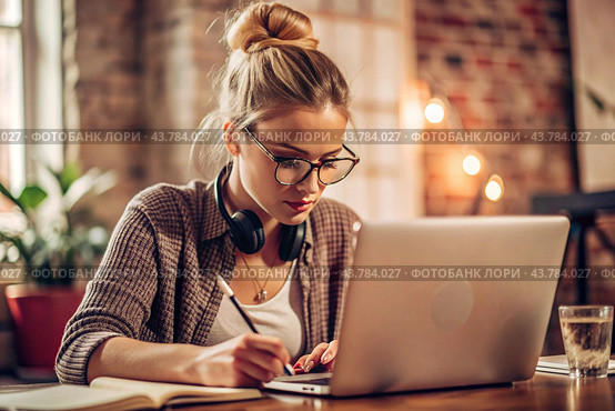 A young female freelance businessman in glasses and headphones is working on a laptop or shopping in an online store. The woman is writing something in a notebook. Стоковое AI-изображение, AI-художник Photojuli / Фотобанк Лори