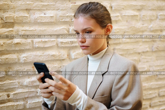 A young woman in a stylish blazer stands confidently against a textured wall, focusing on her smartphone, embodying the essence of modern lifestyle and technology in todays fastpaced world. A focused young woman is fully engaged with her smartphone while in a modern and vibrant setting. Стоковое фото, агентство Ingram Publishing / Фотобанк Лори