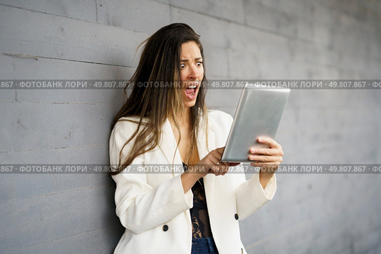 A young woman wearing a stylish white blazer is expressing shock and disbelief while intently looking at her tablet, clearly showcasing a mix of strong emotions against a muted gray background. A shocked woman is reacting to breaking news she is reading on her tablet against a gray wall. Стоковое фото, агентство Ingram Publishing / Фотобанк Лори