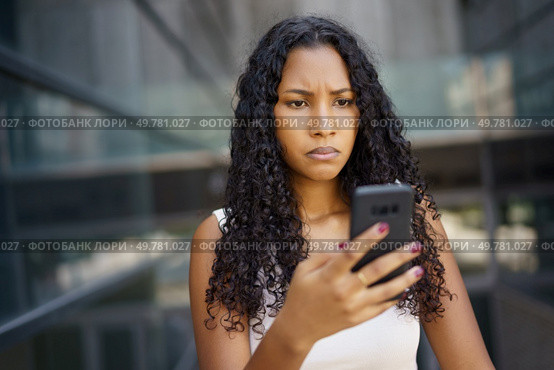 A young woman with curly hair looks worried while checking her smartphone in a busy urban setting, showcasing how modern technology affects our emotions and social interactions daily. A Concerned Woman is Interacting Thoughtfully with Her Smartphone in an Urban Setting. Стоковое фото, агентство Ingram Publishing / Фотобанк Лори