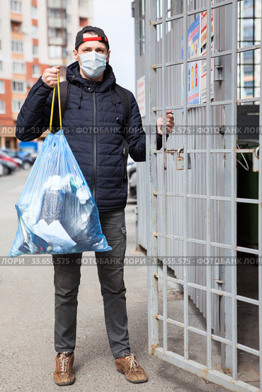 Adult Caucasian man wearing surgical mask taking out the garbage to trash enclosure of residential house. Стоковое фото, фотограф Кекяляйнен Андрей / Фотобанк Лори