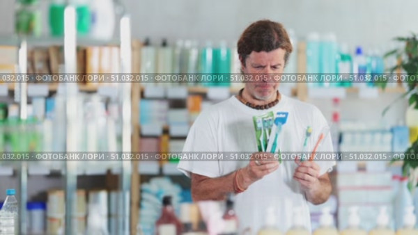 Adult man stands in a European pharmacy and chooses a set of dental care on the recommendation of the dentist. Purchase of dental hygiene products. Стоковое видео, видеограф Яков Филимонов / Фотобанк Лори