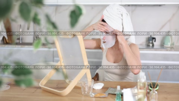 Adult woman in towel turban on head applying hydrating face mask during daily self-care routine after shower at home. Стоковое видео, видеограф Яков Филимонов / Фотобанк Лори
