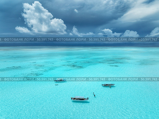 Aerial view of colorful boats and yachts in clear water in summer. Mnemba island, Zanzibar. Top view of sandbank in low tide, blue sea, swimming people, yachts, dramatic sky with clouds. Rainy season. Стоковое фото, агентство Ingram Publishing / Фотобанк Лори