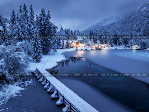 Aerial view of fairy alpine village in snow, bridge, forest, Jasna lake, houses, street lights at winter night. Top view of mountains, illumination, snowy pine trees at dusk. Kranjska Gora, Slovenia. Стоковое фото, агентство Ingram Publishing / Фотобанк Лори