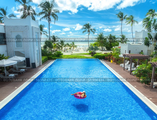 Aerial view of young woman on swimming ring in pool near ocean at sunrise. Top view of girl, blue water, beautiful hotel, palm trees, sandy beach, sea and sky with clouds. Pwani Mchangani, Zanzibar. Стоковое фото, агентство Ingram Publishing / Фотобанк Лори