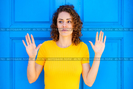 Afraid young female in yellow t shirt with curly brown hair and raised hands looking away against blue wall on street. Scared woman with hands up on blue background. Стоковое фото, агентство Ingram Publishing / Фотобанк Лори