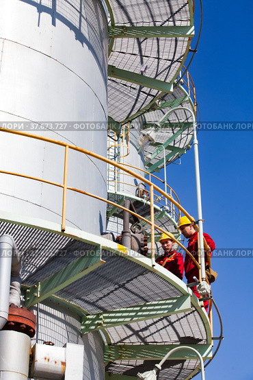 Aktobe region/Kazakhstan - May 04 2012: The grey distillation column of oil refinery plant on blue sky background. CNPC-AMG company. Two maintenance workers in yellow hardhats and red work wear. Редакционное фото, фотограф Alexey Rezvykh / Фотобанк Лори