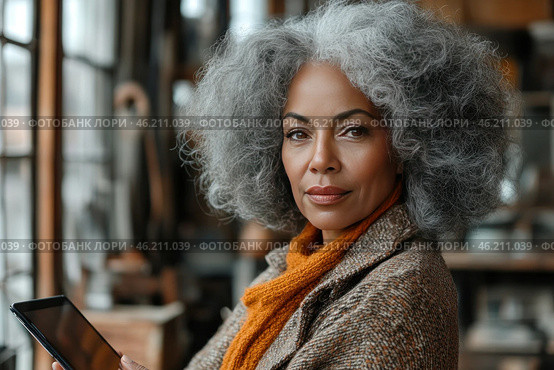 An elegant woman with grey, thick and voluminous curls, sitting in a cozy room with warm natural light. She holds a tablet in her hands and looks with confidence and dignity. Her image is emphasized by a stylish coat and a warm knitted scarf of mustard color. The background is slightly blurred, creating an atmosphere of coziness and concentration, perhaps in a cafe, library or workshop. Стоковое AI-изображение, AI-художник Анна Кривицкая / Фотобанк Лори