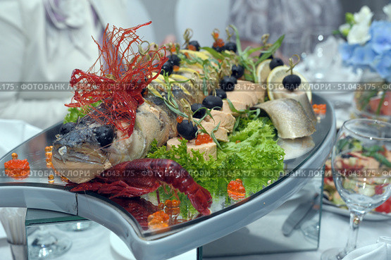 An exquisite festive dish — stuffed and decorated fish served on a large mirrored platter. The head of the fish is decorated with an edible element of bright red caramelized sugar, and the eyes are decorated with olives. Around the fish are pieces of fillet, lemon rolls, olives on skewers, fresh herbs, lettuce leaves and red caviar. In front, near the head of the fish, lies a boiled bright red crayfish, adding to the effect of the composition. The garnish and decor are arranged symmetrically, creating a festive and luxurious look. The dish is served at a banquet, as evidenced by the glasses, salads and smartly dressed guests in the background. Стоковое фото, фотограф Анна Кривицкая / Фотобанк Лори