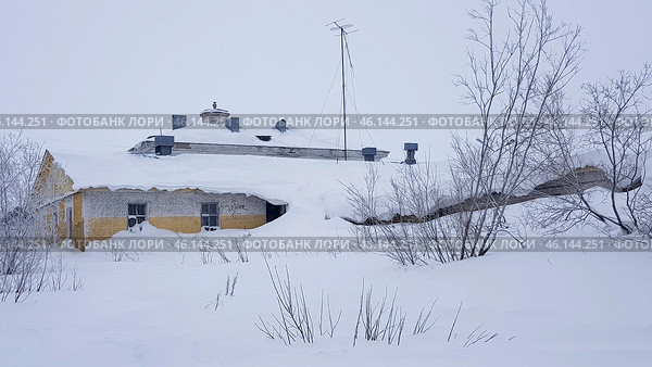 An old snow-covered building in Vorkuta, partially covered with snowdrifts. It is clear that the snow has accumulated up to the first-floor windows, almost completely hiding the lower part of the building. The roof is covered with a thick layer of snow, and dry bushes covered with snow are located nearby.A tall antenna is installed on the roof of the building, which may indicate its past administrative or industrial significance. Perhaps this is an old headquarters, mine administration, or utility facility. The walls of the building are painted yellowish-gray, which contrasts with the whiteness of the snow and the gray sky.Judging by the condition of the windows, facade, and snowdrifts, the building is either abandoned or used very rarely. The photograph perfectly conveys the atmosphere of the harsh polar winter in Vorkuta. Стоковое фото, фотограф Анна Кривицкая / Фотобанк Лори