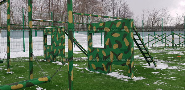 An outdoor sports ground with obstacle course elements. In the center of the frame are two massive metal and wood structures, painted green with a camouflage pattern. Each structure has square holes cut out to imitate windows, and on the side are ladders for climbing. These elements are intended for obstacle course training, physical training, or military sports. In the foreground are visible chains and bars for exercises, and in the background are other elements of the obstacle course. The ground is covered with artificial turf with remnants of snow. The background is a metal fence and bare winter trees. The atmosphere of the picture is emphasized by the combination of outdoor sports, winter season, and the military style of the exercise equipment. Стоковое фото, фотограф Анна Кривицкая / Фотобанк Лори