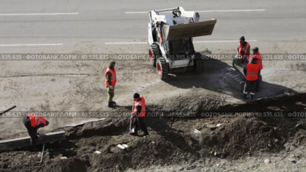 ANGARSK, IRKUTSK REGION, RUSSIA - May 18, 2023: Video of workers in signal red vests lay curb stone on the side of road being repaired. Mini tractor loader brings soil in bucket. Top view. Редакционное видео, видеограф Виктория Катьянова / Фотобанк Лори