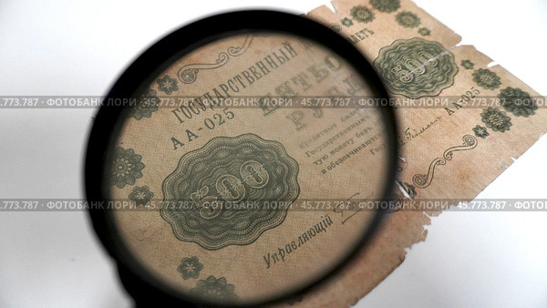 Антикварная банкнота 500 рублей 1918 года под лупой.  Фрагмент старинной купюры номиналом 500 рублей, датированной 1918 годом, рассматриваемой через увеличительное стекло. Видны детали оформления, включая орнаменты, номинал, серийный номер и текст о гарантиях обеспечения золотом. Банкнота имеет повреждения и следы износа, указывающие на её историческую ценность. Стоковое фото, фотограф Анна Чижова / Фотобанк Лори