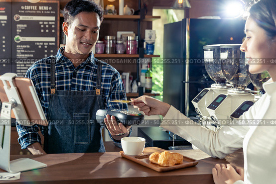 Asian customer using her credit card with contactless nfs technology to pay a barista for her coffee purchase at a cafe bar. Стоковое фото, агентство Ingram Publishing / Фотобанк Лори
