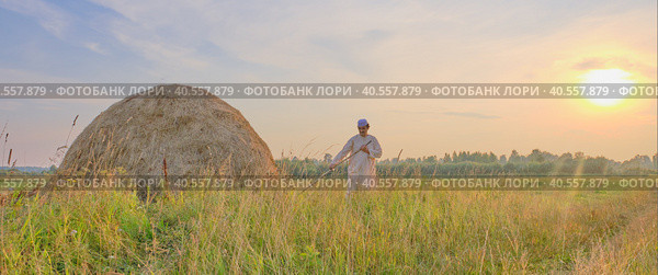 At sunset an senior Muslim man in an embroidered skullcap and white traditional Clothes mows hand-scythe grass in a hayfield. Стоковое фото, фотограф Ирина Кононова / Фотобанк Лори