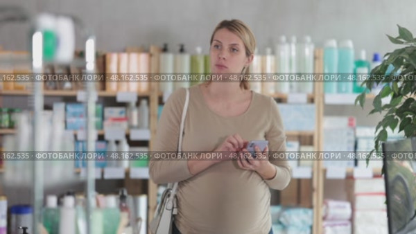 Attentive pregnant woman scrolling phone while looking for something in drugstore. Стоковое видео, видеограф Яков Филимонов / Фотобанк Лори