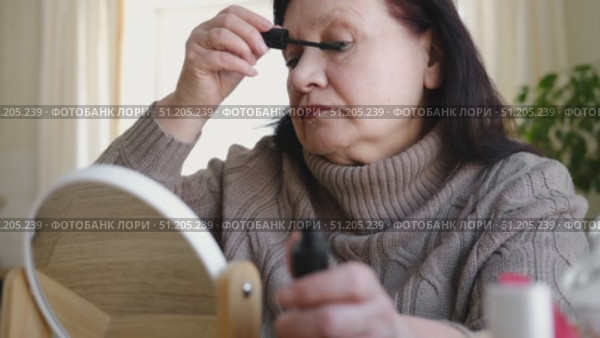 Attractive brunette senior woman putting on makeup looking in the mirror. Beautiful lady caring her face. Elderly female applied cosmetics. Natural old beauty and aging concept. Skin care and make-up. Стоковое видео, видеограф Марина Демидюк / Фотобанк Лори