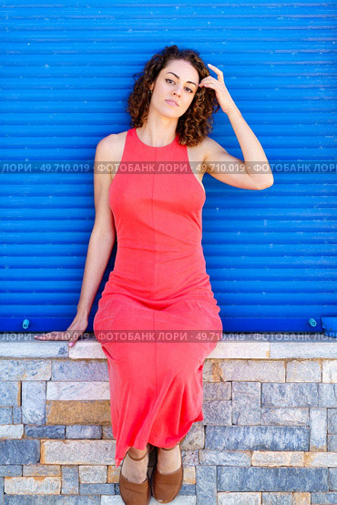Attractive young female in red sundress, sitting on stone wall with sandals and looking at camera while touching hair with hand in daylight against blue shutters. Young woman sitting on wall in red dress. Стоковое фото, агентство Ingram Publishing / Фотобанк Лори