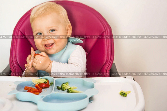 Baby food. The first meal. Feeding the baby. A cute little kid eats vegetables. Healthy food. The child is sitting on a high chair for feeding. Fresh vegetables and fruits. Стоковое фото, фотограф Photojuli / Фотобанк Лори
